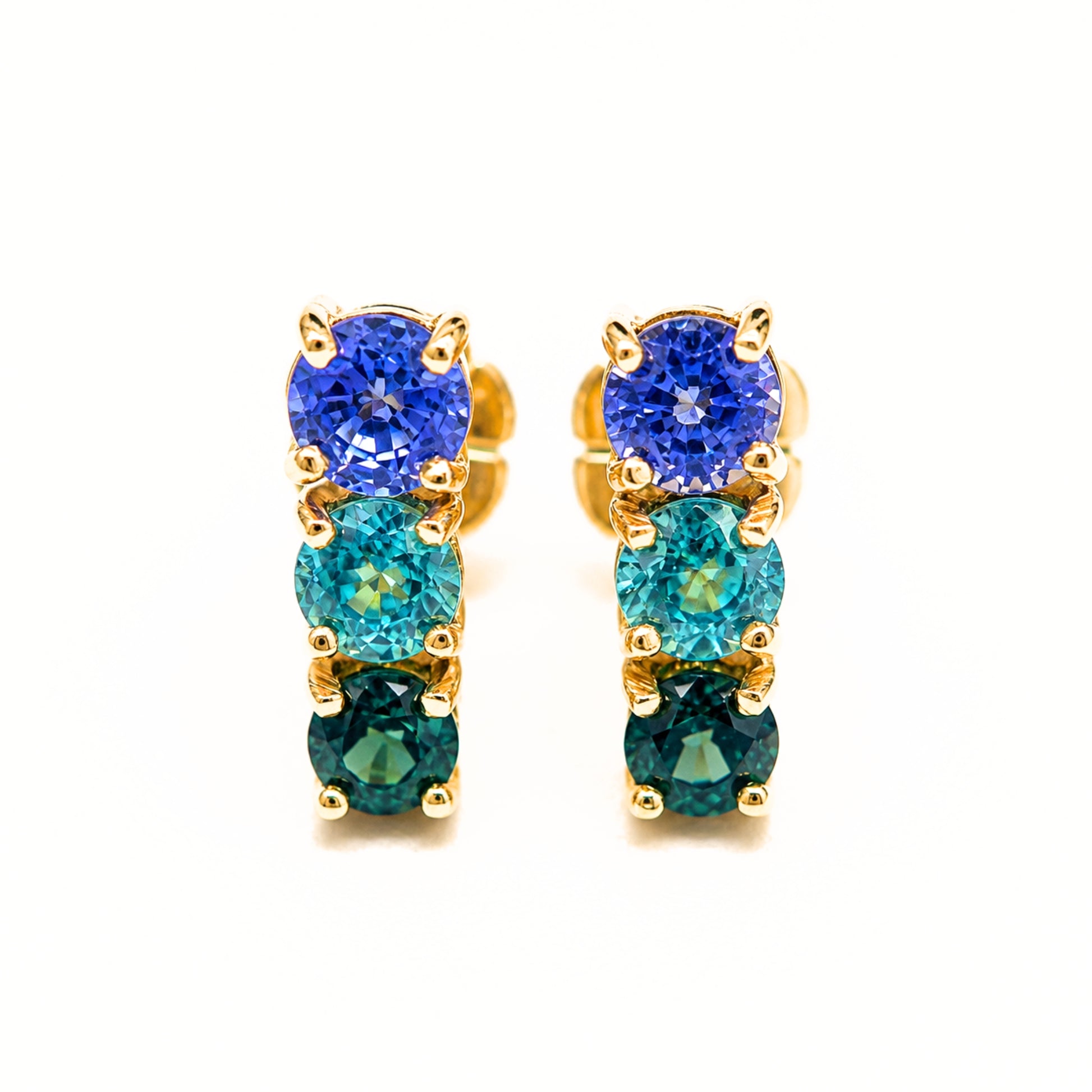 A pair of gold drop earrings featuring three vertical rows of round-cut gemstones in shades of royal blue, teal, and emerald green, set in yellow gold prong settings against a plain white background.
