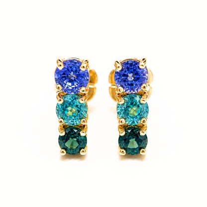 A pair of gold drop earrings featuring three vertical rows of round-cut gemstones in shades of royal blue, teal, and emerald green, set in yellow gold prong settings against a plain white background.