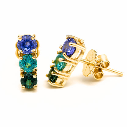 A pair of gold stud earrings featuring three vertical rows of blue sapphire, blue zircon, and green sapphire shown on a clean white background with one earring facing forward and the other angled to show the post and butterfly back.