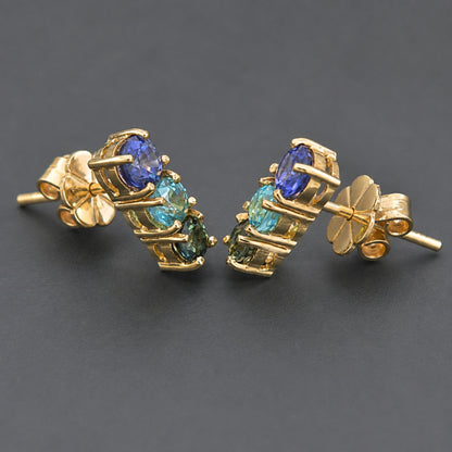 A pair of gold post earrings with a cluster of round-cut blue sapphire, blue zircon, green sapphire, displayed on a dark gray background.