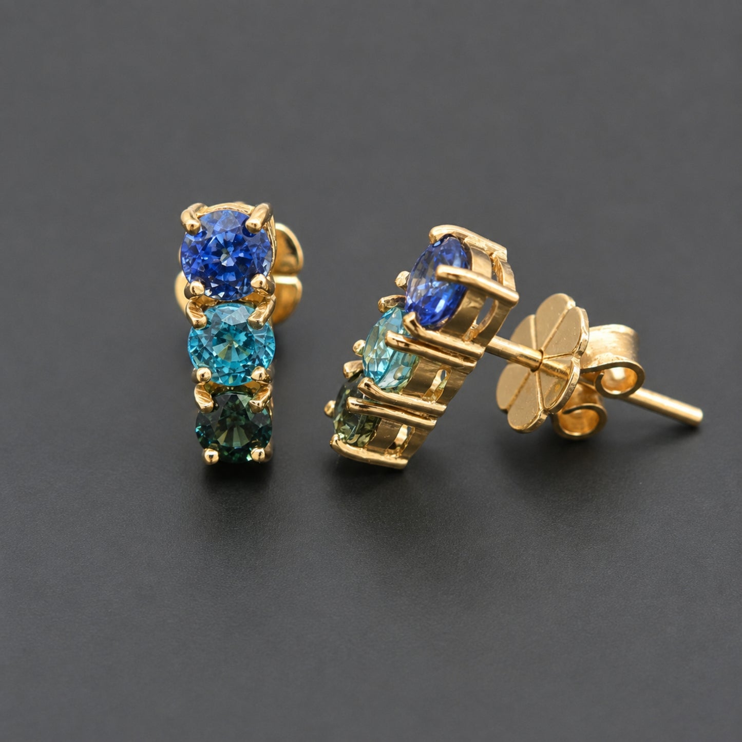 A pair of gold stud earrings featuring three round-cut gemstones in a vertical stack, with blue, teal, and emerald green stones set in a gold prong setting. One earring is shown from the front and the other from the side/back, revealing the butterfly clutch post, on a dark gray background.