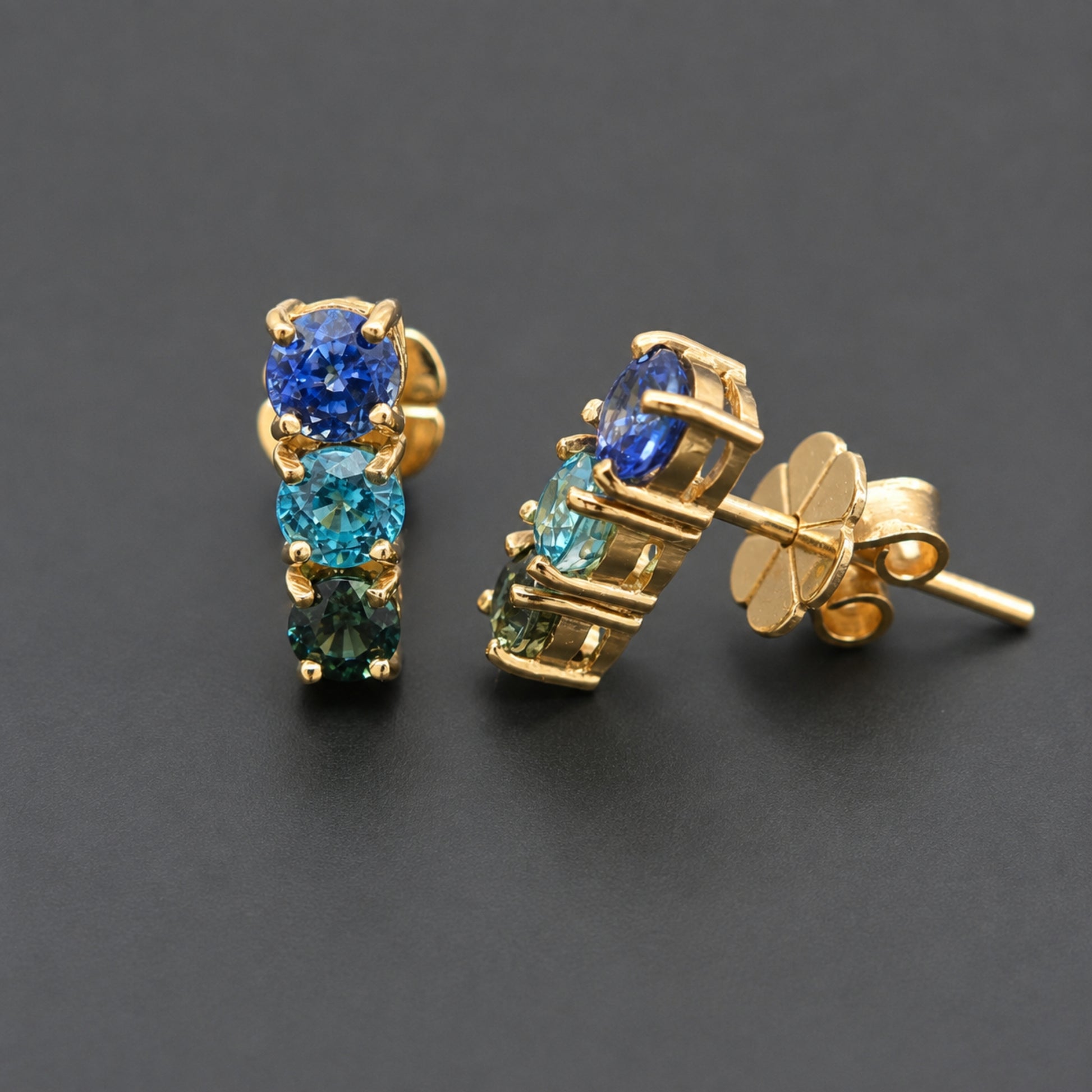 A pair of gold stud earrings featuring three round-cut gemstones in a vertical stack, with blue, teal, and emerald green stones set in a gold prong setting. One earring is shown from the front and the other from the side/back, revealing the butterfly clutch post, on a dark gray background.