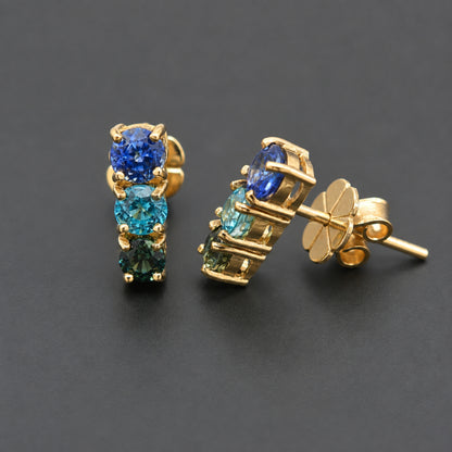 A pair of gold stud earrings featuring three round-cut gemstones in a vertical stack, with blue, teal, and emerald green stones set in a gold prong setting. One earring is shown from the front and the other from the side/back, revealing the butterfly clutch post, on a dark gray background.